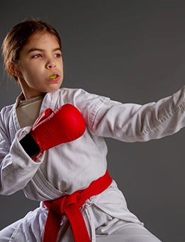 A little girl wears a white robe, red belt, and red boxing gloves while sporting a mouthguard to protect her smile